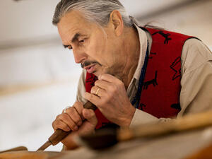 Man with gray hair and bright colored vest carves an unseen wooden object.