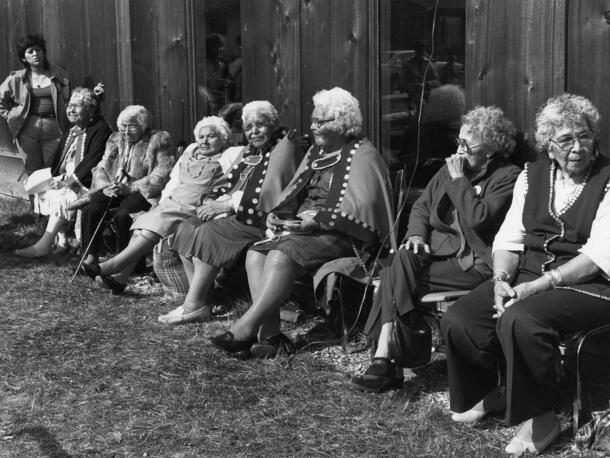 7 older women sit on chairs in a row outside a building, with an 8th woman standing on left end of line.