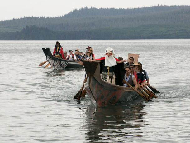 Two canoes full of people on the water. In one, a person in an eagle mask stands at front and in the other, a person in a robe stands at front. 