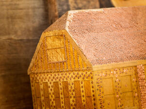 Carved piece shaped like a building covered in hammered down copper nails on the roof and in lines against the sides.