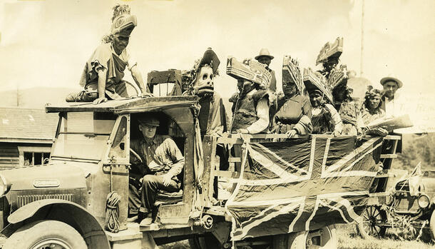 Seven people wearing headdresses stand in the back of an early-1900s pickup truck.  A man with a hat holds a hand-written sign. 