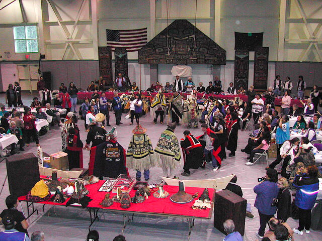 In the center of a large room, a number of people wearing ceremonial regalia stand in a circle with a drummer while spectators look on.