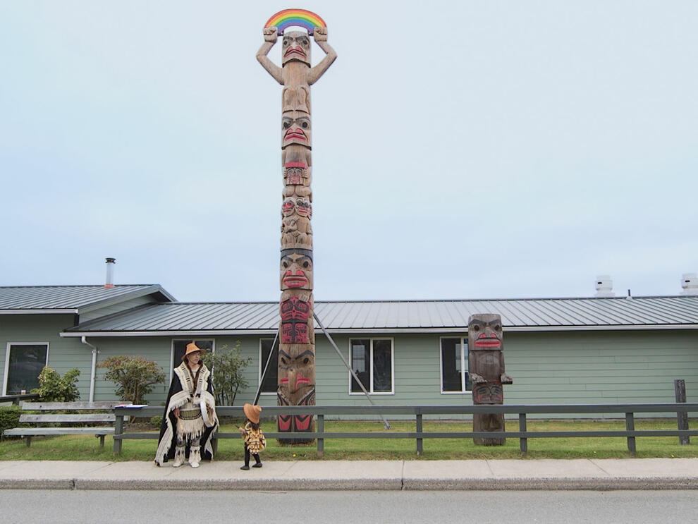 Man in fringed boots, skirt, robe and hat and small child in big hat stand in front of memorial pole, which is on the lawn of a low house.