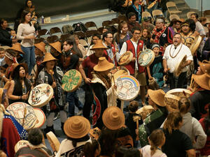 Large group of people in a room stand in a circle, many wearing straw hats and holding decorated drums, as they drum, sing, and dance.