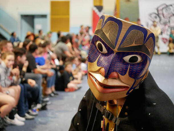 Close-up of a person dancing in a large wooden mask with painted features and patterns, as a blurry crowd in the left-hand background watch.