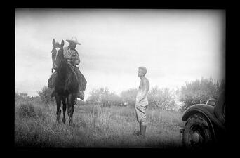 Alfred Kinsey stands in a grassy field and talks to a man in a large hat on horseback, with the front wheel of a car visible in the right foreground.
