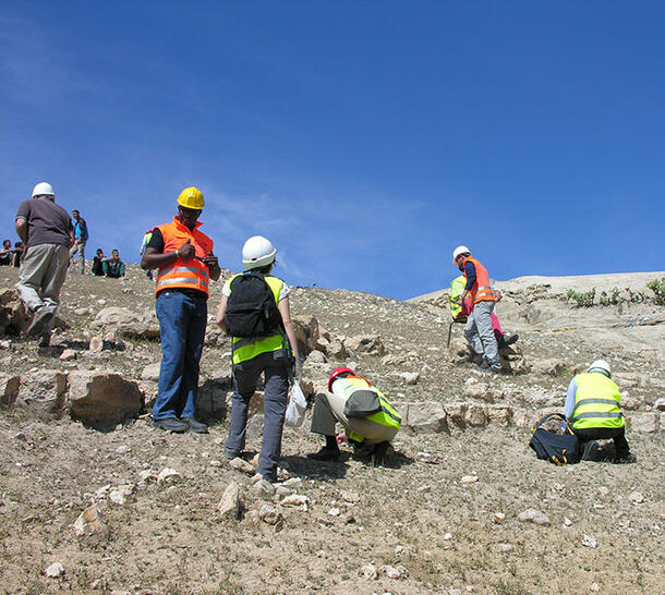 Seven people in the foreground wearing hard hats and most wearing bright vests stand on rocky hill, with 5 more people sitting in the background.