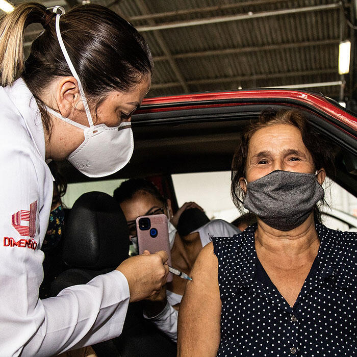 Woman seated in the passenger seat of a car receives a vaccination from a healthcare worker, and the driver of the car takes a cellphone photo.