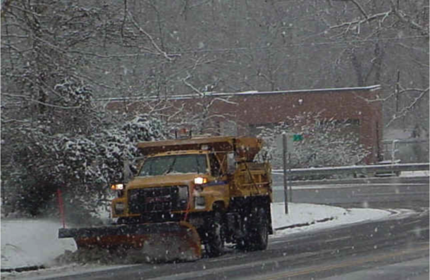 Yellow dump truck fitted with a plow clears a paved road of falling wet snow. A small low building is in background.
