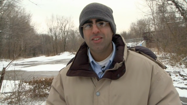A man, scientist Sujay Kaushal, wearing a knit hat, jacket, and lavalier microphone on shirt collar outside in the snow, with trees in the background.