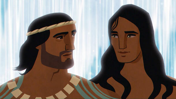 A Hawaiian warrior and a handsome young man stand side-by-side, looking at one another, as they stand in front of a waterfall.