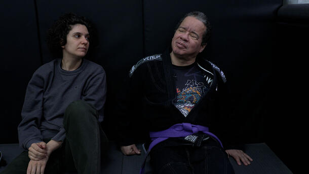 Angel Adorno, a blind Brazilian jiu-jitsu artist, wears their belted robe and sits beside another person against a wall.