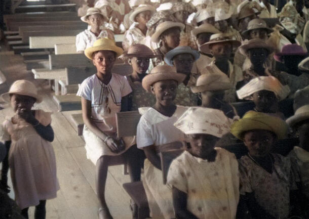 Over twenty young children sit in rows, all wearing brimmed hats and dresses.