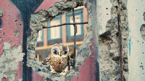 A chipped porcelain figurine of an owl perched in an uneven hole in a wall partially destroyed by bombardment.
