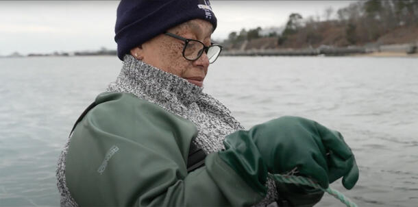A person from the Shinnecock Nation wearing a beanie, scarf and glasses stands on a boat with the water visible in background.