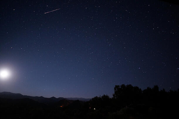 A nighttime landscape shows the glow of the moon and a meteor streaking through the night sky.
