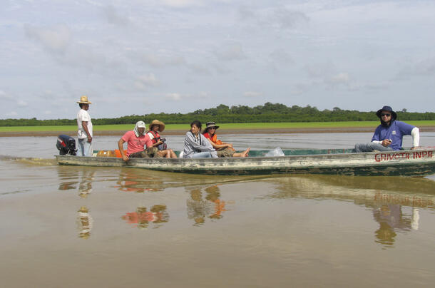 Five people are seated in a low long boat, and one person stands at the motor guiding the boat along the river.
