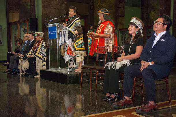 Person in ceremonial dress stands at podium and speaks while several other people  in ceremonial and regular dress sit on either side.