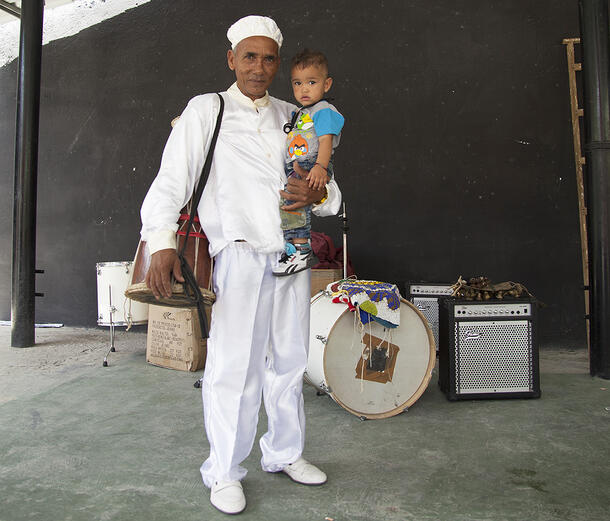 Francisco stands in front of bass drum and other musical equipment. He wears a white cap, shirt, pants and shoes, holds a toddler-aged child.