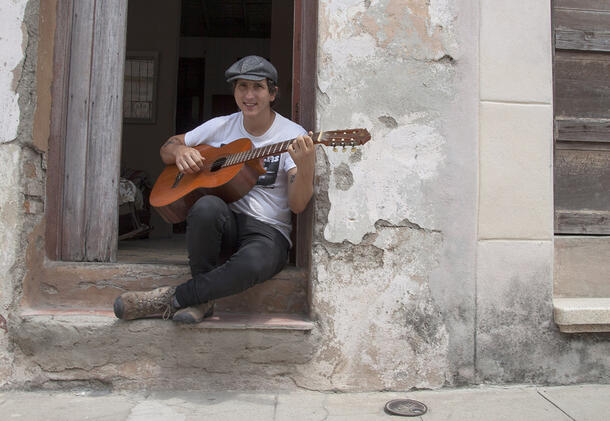 Pablo sits outside on the steps leading to the entrance of his building and plays an acoustic guitar.