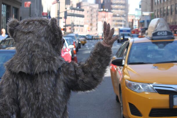 A photo of person seen from behind in a furry animal costume hailing a yellow taxi on a city street.