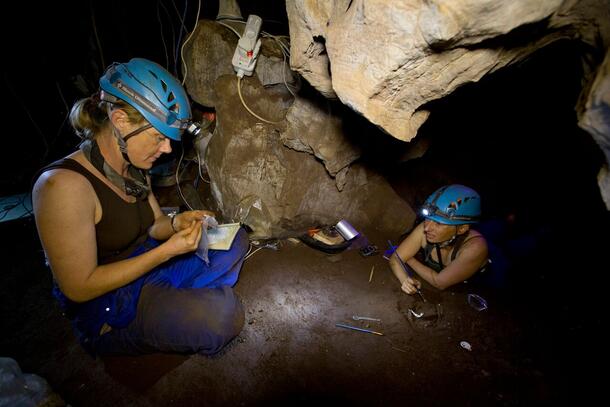 Two women in rock cave wearing helmets with headlamps working with small thin tools.