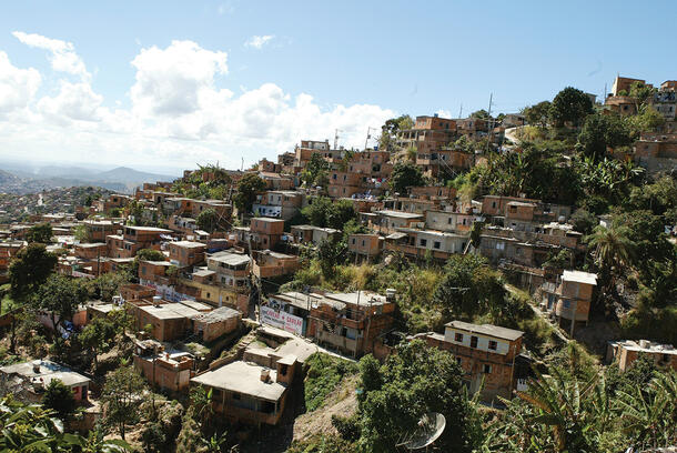 Overhead shot of many small, rectangular buildings on a hill, all surrounded by greenery with an expansive city and mountains visible in background.