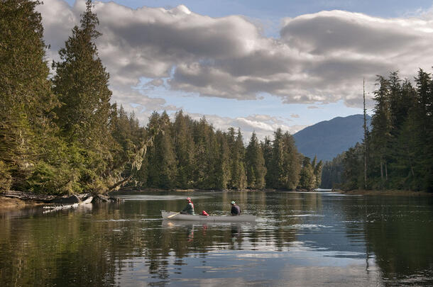 Two people paddle a canoe on an estuary lined with evergreen trees, mountain peaks visible in the background.