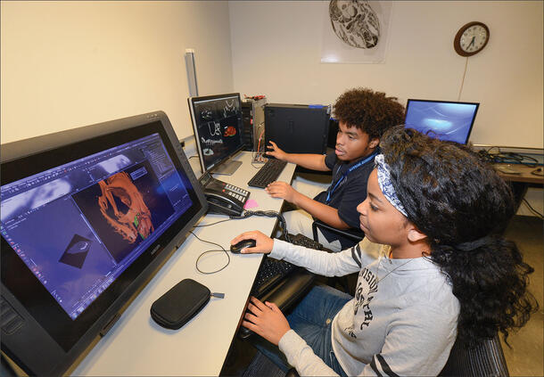 Students Johanna and Xavier sit side-by-side in front of computer monitors displaying CT scans.