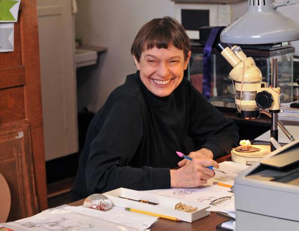 Woman seated at office desk, smiling at camera, holding pen. Several drawings, writing tools, a decorative paperweight, and a microscope are on desk.