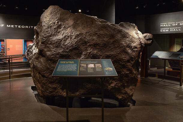The Ahnighito meteorite. A thirty-four ton mass of iron with a rough bumpy pitted surface. In an exhibition hall with explanation panel in foreground.