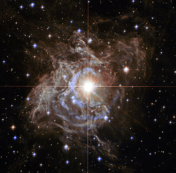 In a nebula, against the inky black of outer space dotted with stars, the RS Puppis, a Cephid variable star appears as a large bright spot.