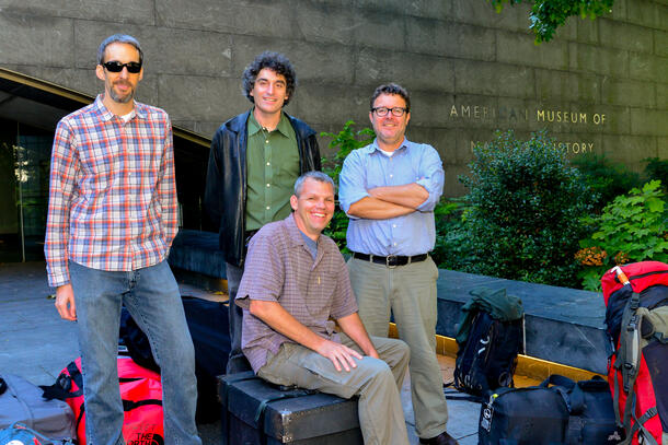 A photo of four men posing outside the American Museum of Natural History with backpacks and gear bags strewn around them and one man sitting atop a large luggage case.