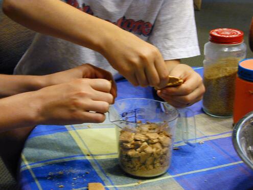 Two pairs of boys' hands breaking up graham crackers over a measuring cup that is half-filled with other broken pieces. 