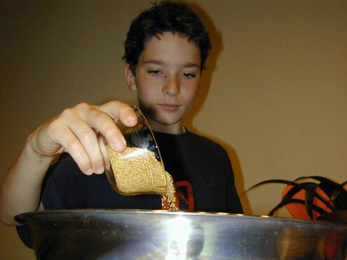 A boy pouring graham cracker crumbs from a small glass cup into a large metal mixing bowl. 