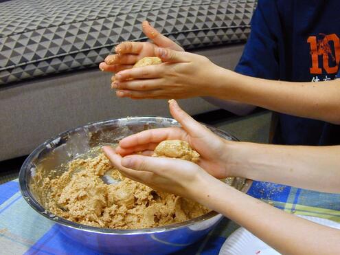 Closeup of two pairs of boys' hands rolling cookie dough into balls before a large metal bowl. 