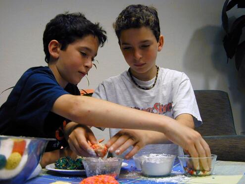 Two boys rolling cookie dough balls in containers of sprinkles and powdered sugar. 