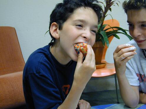 Boy gleefully biting into a decorated cookie ball while his friend—who is also holding a cookie—looks on in the background.