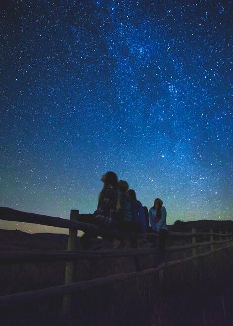 Four girls looking up at the Milky Way in the night sky and sitting on a long wooden fence in a field of tall grass. 