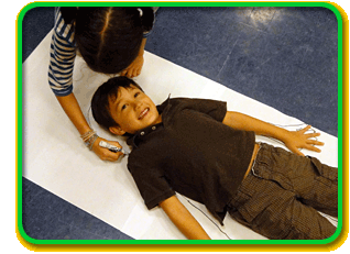 A child laying on a large piece of paper is outlined in marker by another child.