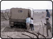 Two people pushing a covered truck in the Gobi desert, with two more people trailing behind.