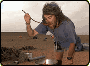 A person with long hair tastes food from a large pot, using a long spoon, with the desert in the background.