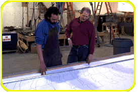 Two people look over a life-sized line illustration of a T. Rex's tail bones which is laid out on a large table.