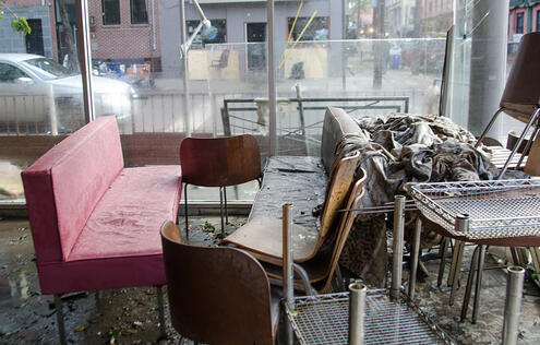 room containing water-damaged furniture. It is on ground level, and, through a partly-damaged plexiglass wall, a street, a sidewalk, and nearby damaged buildings are visible.