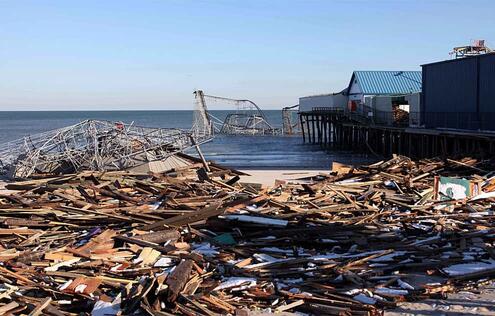 A wide pile of wood planks covering a beach, a damaged beachfront building on stilts, and partly submerged roller coaster in Seaside Heights, New Jers