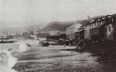 Black and white photo of waves hitting the coast of a town, with people gathered outside.