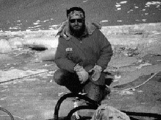 A man, astrobiologist Chris McKay, wearing sun goggles and cold weather gear, kneeling outdoors on a frozen rugged icy surface.