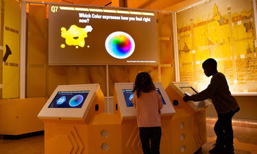 Two children touch the screens of digital kiosks facing a large presentation monitor that asks "Which color expresses how you feel right now?"