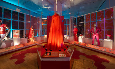 Wide shot of a room featuring displays including a red football uniform, a pink suit, a red sari, and a red chiffon and organza gown in the center