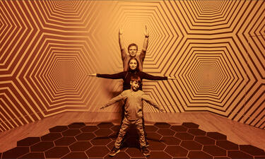 A man, woman, and child stand with arms outstretched in a room with geometric patterned wallpaper that appears colorless.